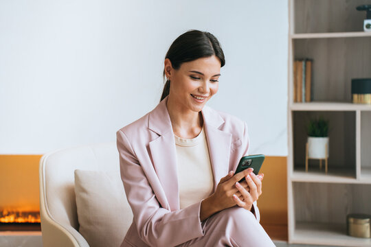 Relaxed with a smile, the woman enjoys her phone in a chic pastel suit, embracing technology in a stylish setting.
