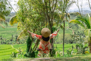 Woman on the swing at the green rice fields wearing long red and white dress and Asian conical hat in Bali Indonesia