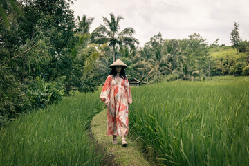 Woman walking along the green rice fields wearing long red and white dress and Asian conical hat in Bali IndonesiaIndonesia