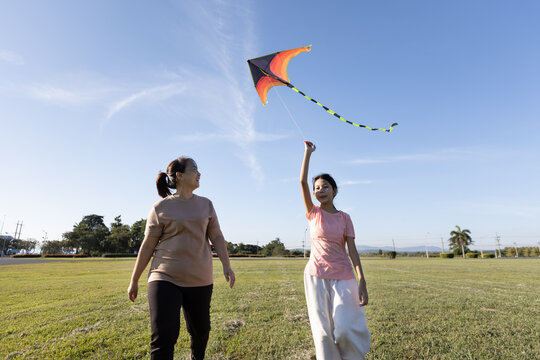 Asian mother and daughter walking together outdoors while flying a kite, showing family bonding, relaxed outdoor activity, and happy childhood moments.