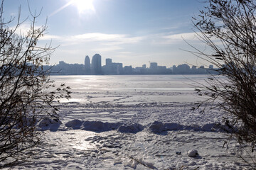 Snow-covered riverbank in the foreground with a large city skyline across a wide frozen river. Bright winter day with clear sky, icy river surface and urban winter landscape