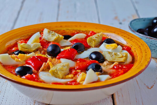 Plato tradicional de ensalada de pimientos rojos asados con huevo duro, at&uacute;n, aceitunas negras y corazones de alcachofa en cuenco de cer&aacute;mica