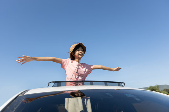 Asian girl with arms outstretched standing through a car sunroof during outdoor travel, showing childhood joy, freedom, and relaxed travel lifestyle.