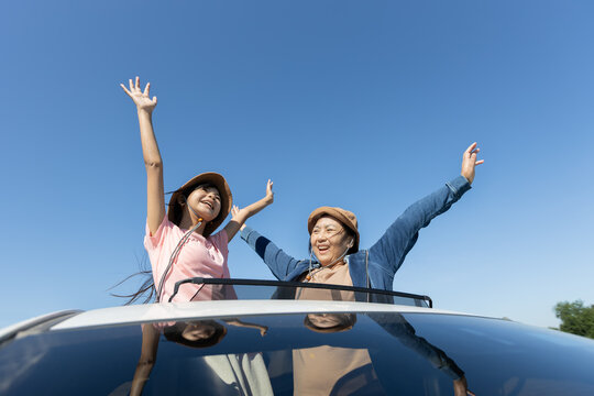 Asian mother and daughter with arms outstretched standing through a car sunroof during travel, showing family joy, freedom, and relaxed outdoor lifestyle.