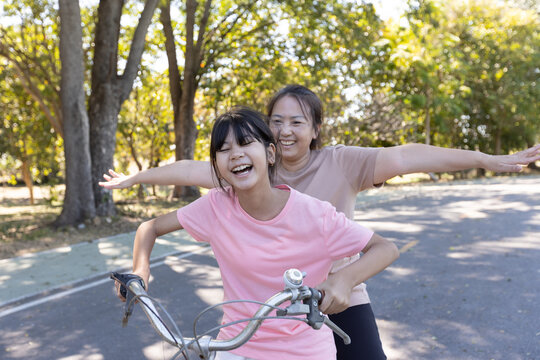 Asian mother and daughter cycling outdoors with arms outstretched and laughing together, showing playful family interaction and joyful lifestyle moment.