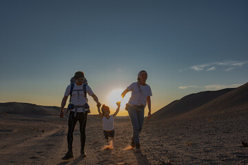 A young family holding their child's hands walking in the desert during golden hour, kicking up...
