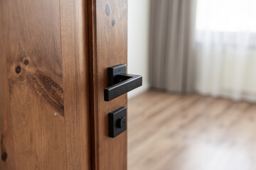 Close-up of a wooden door with a modern matte black handle, partially open to reveal a bright room with wood flooring and sheer curtains