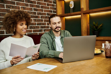 An African American woman and a man sitting at a table with a laptop, engaging in a digital collaboration session.