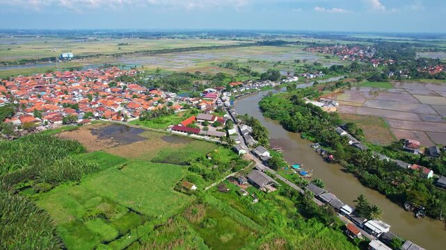 Aerial drone footage of a main brown river, with green wide rice fields and natural fish farms around, with a local village,  fishing boats, in Domas area, Serang regency, Java island, Indonesia