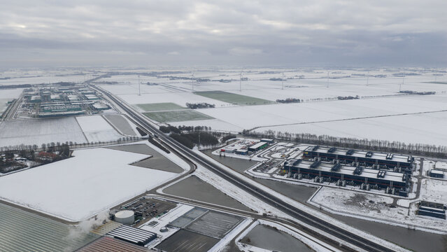 Aerial view of a monochrome winter landscape where snow blankets the flat fields and buildings, contrasting with the dark roads and industrial structures, Middenmeer, North Holland, Netherlands.