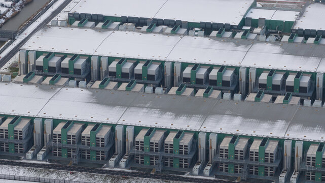 Aerial view of the vast, snow-dusted data center, its metallic structures contrasting sharply with the stark white landscape, Middenmeer, North Holland, Netherlands.