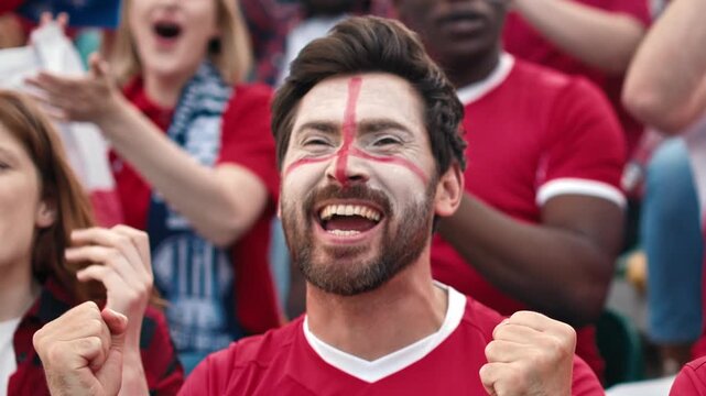 Worried Caucasian male football fan with England flag face paint clasping hands watching intense match. Handsome man in red jersey anxiously reacting in stadium crowd. Outdoors. Close up.