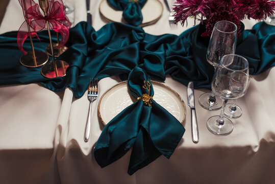 Luxurious banquet table setting with beige tablecloth, green napkins in gold holders, red candles in golden candlesticks and ceramic plates. Refined festive dining decor.