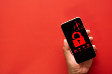 Hand holding smartphone showing red padlock and warning triangle icon on red background. Cyber security breach, system hacked, malware virus, and access denied concept.