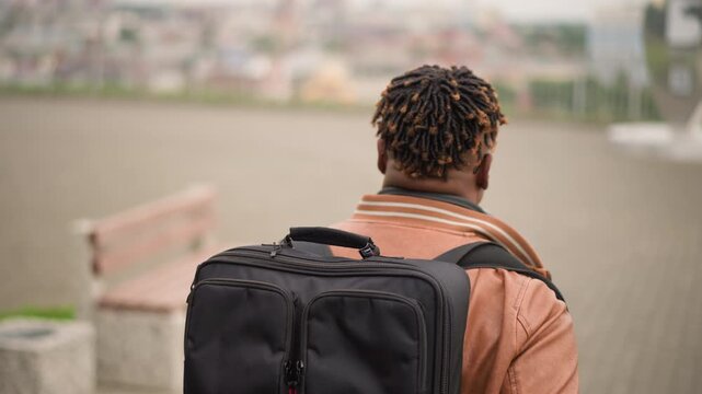 Black man with backpack in plaza walking away from camera, wearing headphones and jacket, rear view closeup with blurred city skyline and empty bench, overcast light creating moody yet hopeful