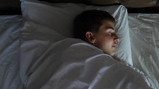 Top view camera moves above a cute teenage boy sleeping in bed with white bedding and smiling happily in his sleep. Comfortable rest on an orthopedic mattress at home.