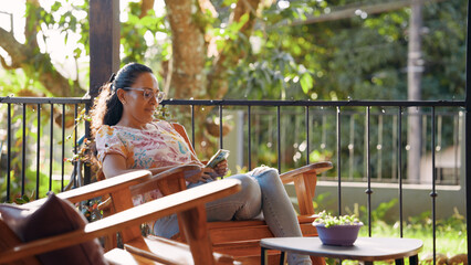 Mature latina woman relaxing on a porch or balcony, sitting in a wooden chair, enjoying sunlight while using a smartphone