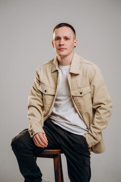 Studio portrait of a young male model sitting on a wooden chair and looking at the camera. Casual outfit with beige cotton shirt, white t-shirt and black cargo pants, isolated background.