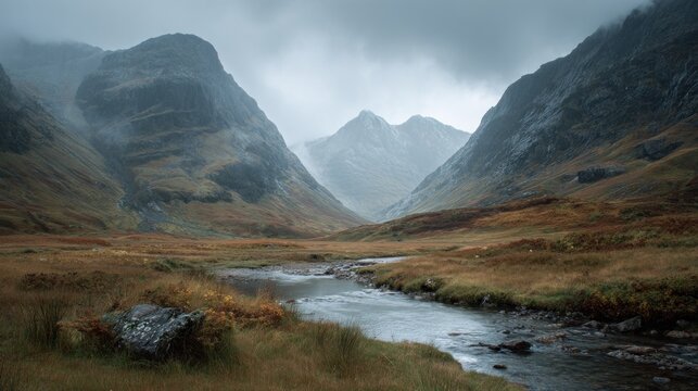 A stream flows through a valley in Glen Coe, Scotland. Mountains rise on either side under a cloudy, misty sky in the Scottish Highlands