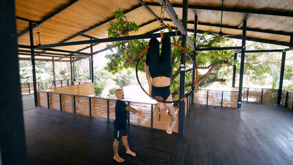 Female aerial dancer hanging upside down on a lyra hoop while an instructor observes, practicing acrobatics and fitness