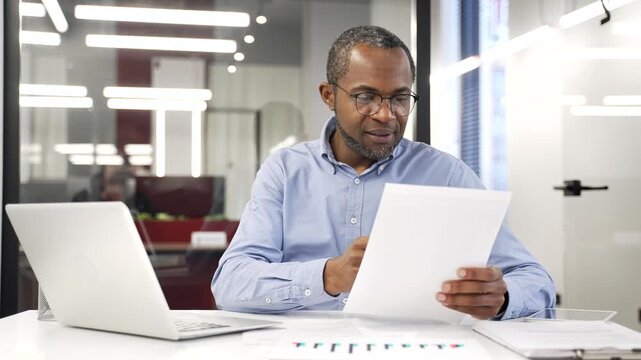 African american businessman is satisfied with financial results looking at document and laptop sitting in a business office. Smiling entrepreneur is happy with positive indicators looking at camera