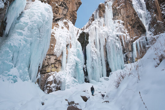 Frozen Butakov waterfall in Almaty, Kazakhstan, with a lone human figure in the background. The powerful blue ice columns of the waterfall contrast with the brown rocks 