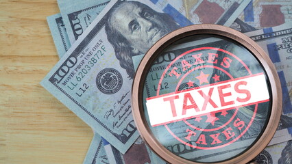 Magnifying glass over US dollar banknotes with taxes stamp representing tax payment, financial obligation, government revenue, audit process, accounting compliance, and taxation concept.