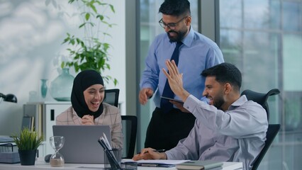 Business Arabian team three Indian colleagues in office discuss problem documents looking at computer happy celebrate work winning achieve looking at screen analyze financial growth success high five