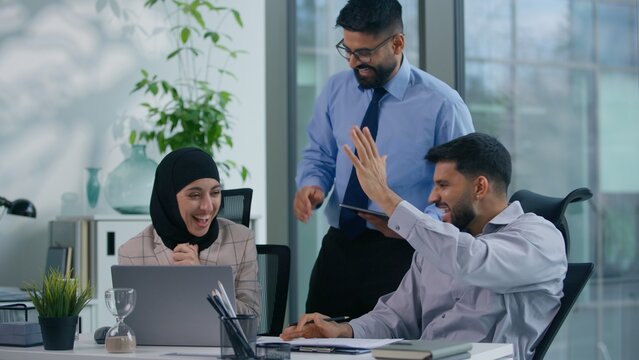 Business Arabian team three Indian colleagues in office discuss problem documents looking at computer happy celebrate work winning achieve looking at screen analyze financial growth success high five