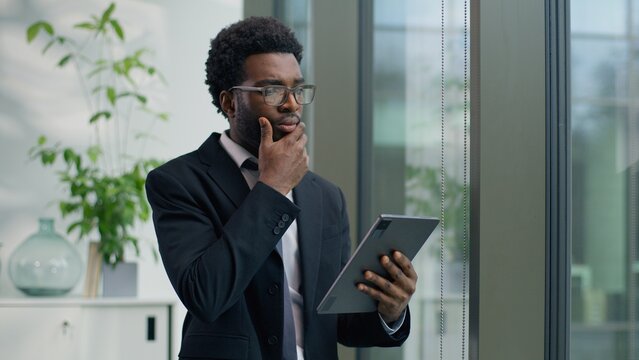 Thoughtful African American businessman in office concentrate using digital tablet looking out window focused business male man guy using technology device thinking planning strategy professional work