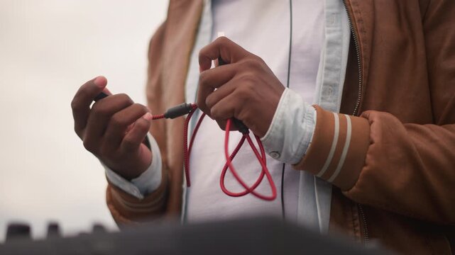 black sound engineer linking red cable into mixer on rooftop, methodical hands threading jack, overcast skyline and urban backdrop, warm jacket sleeves, tactile checks before live outdoor set