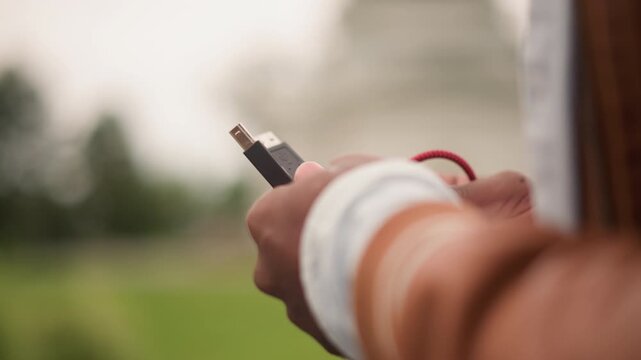 closeup hands holding usb stick in park, fingers adjusting red braided cable, blurred historic building behind, warm film tones, wristband and jacket sleeve visible, quiet anticipation before outdoor