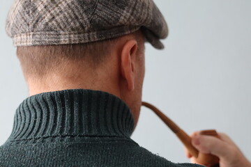 Person in a gray cap holds a wooden pipe and looks away from the camera in a simple indoor setting