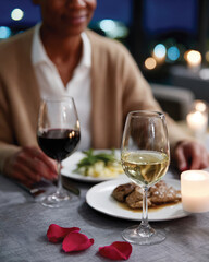 Obraz premium A cozy dining experience featuring a woman holding a glass of white wine, with a delicious meal on the table, illuminated by warm candlelight and romantic rose petals.