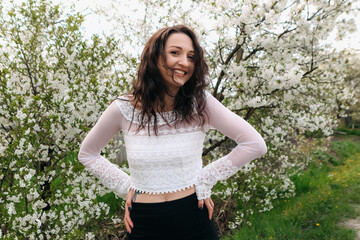 Happy young woman with long brown hair smiling while wearing a stylish white lace top and a chic black skirt, enjoying the beauty of a blooming tree in a sunlit spring garden