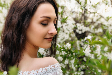 Brunette woman with long, wavy hair wearing a flowing white dress, posing gracefully near a blooming tree in a vibrant spring garden, embracing the serenity and beauty of nature