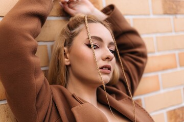 Young blonde woman with braids, wearing a chic brown coat, posing confidently with arms raised against a textured brick wall, exuding a trendy and stylish vibe in an urban setting