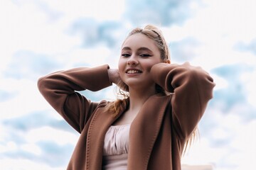 Cheerful blonde woman in a brown coat holds her hands behind her head, enjoying the fresh air and beautiful cloudy sky, showcasing a carefree and happy attitude