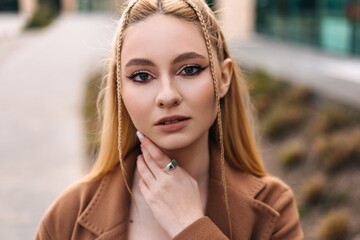 Professional blonde woman in chic brown coat, braided hair framing her face, resting hand thoughtfully on chin while standing against urban background, radiating sophisticated poise