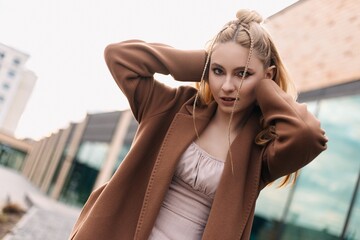 Stylish model exhibiting a trendy braided hairstyle and a chic brown coat, posing confidently with her hands on her head against a backdrop of modern urban architecture