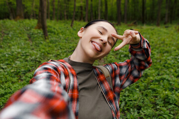 Female hiker sticking out tongue, flashing peace sign while taking selfie amid verdant forest, backpack-wearing adventurer capturing playful moment surrounded by lush greenery