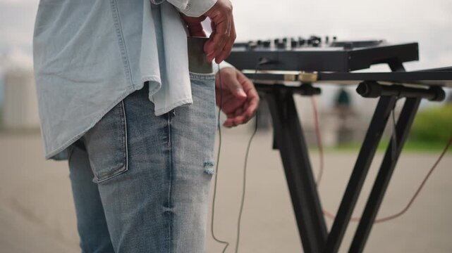 outdoor street dj setting up mixer on urban pavement closeup of hand holding headphones and cables beside compact controller casual denim attire overcast sky anticipatory mood readying for openair