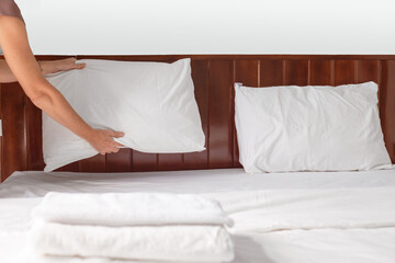 A housekeepers hands fluff and place a white pillow on a neatly made bed with a wooden headboard.