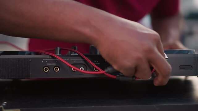 black dj setting up mixer cables on outdoor street table, hands connecting plugs and adjusting knobs, closeup of red wires and fader movements, testing sound through controller and console, gritty