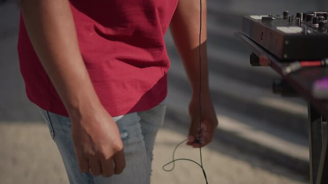 black DJ packing cables into backpack with red interior on sidewalk, unplugging mixer cord and stowing headphones, denim jeans and casual stance, daylight city scene, careful zipper closure, compact