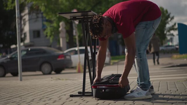 Black musician kneeling to set equipment on pavement, unfolding keyboard stand and pulling mixer from red case, adjusting straps and plugs, preparing streetside performance under warm sunlight