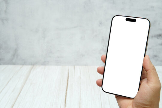 Hand holding smartphone with blank white screen on bright wooden desk and light wall background. Concept of mobile mockup, app presentation, digital marketing, UI UX design, and screen replacement.