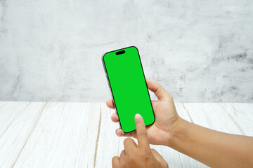 Hand holding smartphone with green screen on bright wooden desk and light wall background. Concept of mobile mockup, app presentation, digital marketing, UI UX design, and screen replacement.