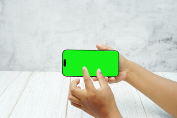 Hand holding smartphone with green screen in landscape orientation on light wall background. Concept of mobile mockup, video ads, app demo, digital marketing, and screen replacement.