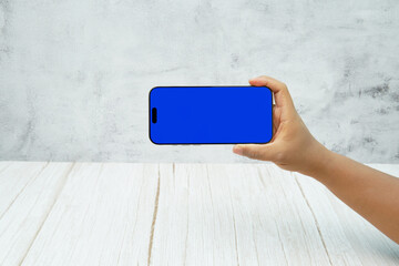 Hand holding smartphone with solid blue screen in landscape orientation on bright wooden desk and light wall background. Concept of mobile mockup, app demo, video advertising, and digital branding.